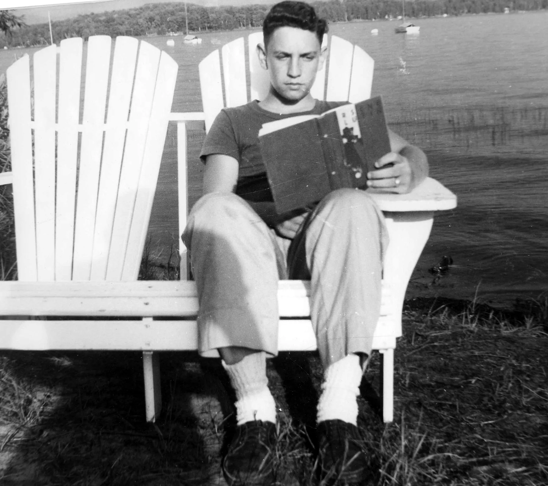 A young man reading in an Adirondack chair by a lake